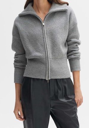 Pull zippé en maille côtelée grise à manches longues, associé à un pantalon en cuir noir taille haute, coupe ample.