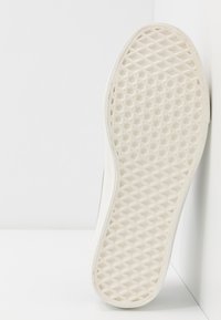Semelle blanche de basket avec motif gaufré appuyée contre un mur blanc, montrant une texture détaillée et un design d’adhérence.