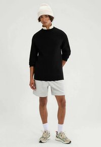 Black ribbed sweater with a high collar, paired with grey shorts. The model wears a white knit beanie and athletic shoes with multi-colored accents.