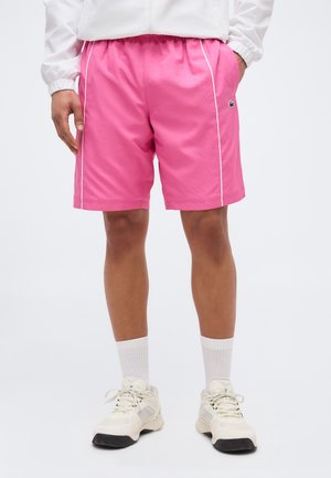 Mann trägt leuchtend pinke Sportshorts mit weißer Paspelierung, weiße kurze Socken und weiße Turnschuhe, steht mit den Händen in den Taschen.