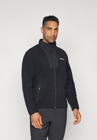 Black fleece jacket with a high collar, contrasting front panel, zippered side pockets, and a logo on the left chest area.