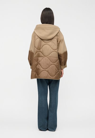 Καπιτονέ καφέ μπουφάν με κουκούλα και στρογγυλές ραφές, συνδυασμένο με χαλαρό μπλε τζιν παντελόνι. Μονόχρωμο σχέδιο με μήκος μέχρι τη μέση.