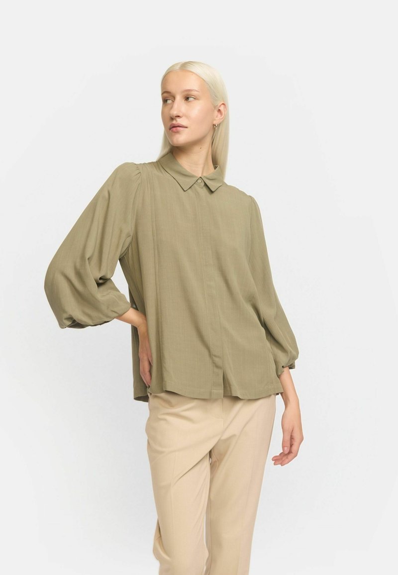Blusa verde oliva con colletto a camicia, maniche lunghe a sbuffo e un taglio morbido. Tessuto liscio con pieghe verticali al centro.