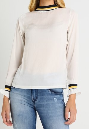 Lichte, doorschijnende witte blouse met lange mouwen, zwart en gele gestreepte kraag en ruches aan de manchetten. Gemaakt in combinatie met blauwe denim jeans.