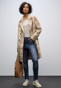 Beżowy płaszcz w kratę z wełny, jasna bluzka, ciemnoniebieskie skinny jeans, torba z jasnego zamszu oraz beżowe sneakersy. Płaszczyk ma podwójne guziki i kieszenie.