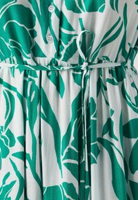 Robe florale verte et blanche avec fermeture à boutons, ceinture à la taille et texture de tissu plissé. Le design met en avant de grands motifs de feuilles.