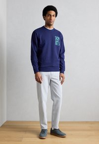 Tmavomodrá mikina s tyrkysovým grafickým potlačou na hrudi, v kombinácii s svetlými rifľovými nohavicami a sivými teniskami s hnedými podrážkami.