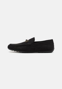 Mocassins en suede noir avec un design surpiqué et un accent de matériel décoratif. Semelle plate avec un motif de grip texturé, silhouette basse.