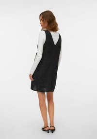 Juodas tekstūruotas suknelė su V formos nugaros iškirpte ir paprasta siluetu, dėvima ant baltos ilgomis rankovėmis palaidinės, užbaigta aukštakulniais.