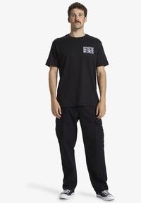 Camiseta de algodón negra de manga corta, con un diseño de bolsillo en olas moradas y verdes; combinada con pantalones cargo negros y zapatillas negras.