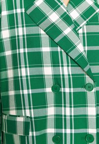Zelený kostkovaný blazer s dvouřadým designem, dvěma zelenými knoflíky a strukturovanou tkaninou s bílými akcenty a různými šířkami kostkování.