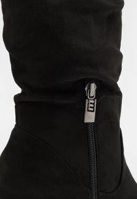 Botte en daim noire avec une finition texturée, dotée d'une fermeture éclair latérale en argent métallique. La partie supérieure est froncée, créant une silhouette décontractée.