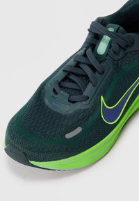 Ciemnozielone i czarne buty do biegania Nike z niebieskim i neonowozielonym swooshem, czarnymi sznurówkami i neonowozieloną podeszwą, oznaczone "Stellar Ride".