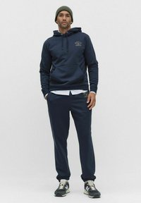 Man i marinblå hoodie, matchande joggerbyxor, grå beanie, vit undertröja och svartvita sneakers står mot en enfärgad bakgrund.