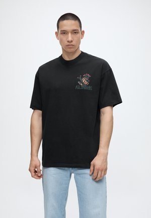 Muž nosiaci čierne oversized tričko s grafickou potlačou na ľavej strane hrude a svetlomodré rifle, stojaci pred jednofarebným svetlým pozadím.