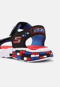 Mustat sandaalit sinisellä sisäpuolella, säädettävällä hihnalla ja monivärisellä, teksturoidulla pohjalla, jossa on punaisia, valkoisia ja sinisiä neliöitä.