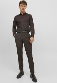 Camicia fitted marrone scuro con colletto button-down abbinata a pantaloni marroni a quadri. Scarpe in pelle nere e una cintura nera completano l'outfit.
