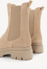 Béžové semišové kotníkové boty mají elastické boční panely, robustní podešev a hladký povrch s výraznými švy a poutky pro přetahování.