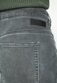 Mörkgrå denimjeans med en texturerad yta, med kontrasterande sömmar och en svart läderlogotyp vid midjan.