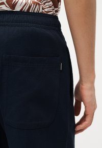 Άτομο που φοράει σκούρο μπλε παντελόνι με πίσω τσέπη και καφέ πουκάμισο με σχέδια, ορατό από τη μέση και κάτω, με το χέρι χαλαρά στο πλάι.