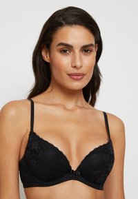 Jeune femme portant un soutien-gorge en dentelle noire avec des bretelles fines, se tenant de face devant un fond clair.