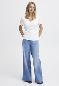 Weißes V-Ausschnitt-T-Shirt mit einem kleinen roten Kirschgrafik, kombiniert mit hellblauen Schlagjeans, die eine ausgewaschene Optik und einen rohen Saum haben.
