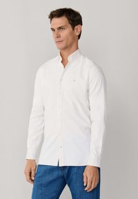 Man die een wit overhemd met lange mouwen en een blauwe spijkerbroek draagt, staand tegen een effen lichtgrijze achtergrond.