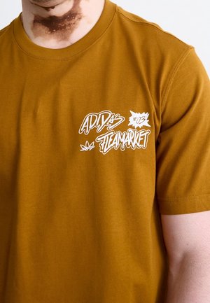 Brązowy t-shirt z białym graficznym tekstem o treści "ADIDAS STEAMARKET No 03", noszony przez osobę z widoczną ciemną plamą skórną na szyi.