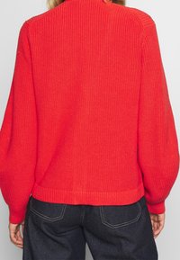 Suéter rojo de punto con cuello y puños acanalados, que presenta mangas de globo y un corte relajado. Tejido texturizado y de color sólido.