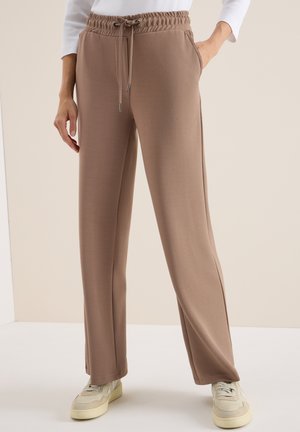 Pantalons de jogging extensibles marron avec une taille élastique, un cordon de serrage, des poches latérales et une coupe décontractée, associés à des baskets de couleur claire.