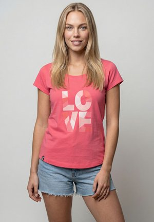 Junge Frau mit langen blonden Haaren, die ein rosa "LOVE"-T-Shirt und blaue Jeansshorts trägt, vor einem schlichten grauen Hintergrund steht.