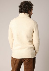 Crème kleurige geribbelde coltrui van zacht materiaal, met een aansluitende snit. Bruine corduroy broek zichtbaar aan de onderkant.
