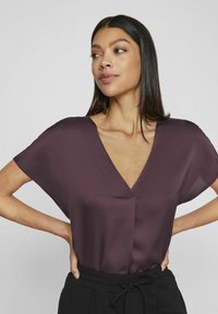 Burgundi spalvos trumpų rankovių bliuska su V formos iškirpte, pagaminta iš lygaus audinio, pasižyminti laisvu siluetu ir subtiliu priekiniu siūlės akcentu.