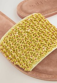 Sandalias de color amarillo con una tira texturizada adornada con pequeños patrones de flores y cuentas rosas, sobre una plantilla de ante color marrón.