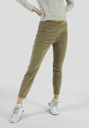 Persoon met olijfgroene ribfluwelen broek, grijze gebreide trui en witte sportschoenen, staand met handen in de zij.