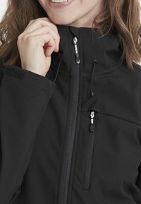 Svart softshell-jacka med hög krage, frontdragkedja, snörning och två zip-fickor; har en slät yta och minimalistisk design.