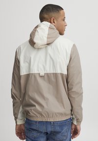 Jachetă ușoară cu glugă în bej și alb-off. Caracteristici un design cu blocuri de culoare, manșete elastice și un tiv strâns. Textură netedă.