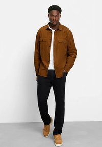 Braunes Hemd mit Knöpfen und zwei Brusttaschen, weißes T-Shirt darunter, schwarze Hose und beige Sneaker. Glatte Textur, klassischer Schnitt.