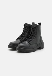 Bottines en cuir noir avec un bout rond, un laçage à l'avant et des détails surpiqués. Semelle plate et texturée avec un léger talon.