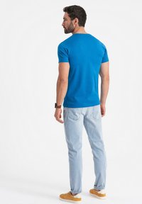 Modré bavlněné tričko s krátkým rukávem, kombinované se světle modrými džíny a žlutými teniskami. Pozadí je jednoduché bílé.