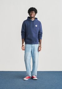 Μπλε σκούρο φούτερ με μπροστινή τσέπη και λογότυπο, συνδυασμένο με ανοιχτό μπλε τζιν και κόκκινα αθλητικά παπούτσια. Απλός σχεδιασμός, casual εφαρμογή και απαλό υφή.