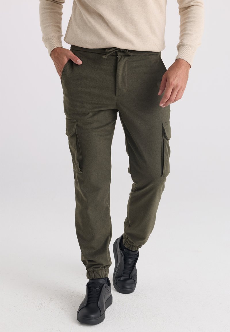 Personne portant un pantalon jogger cargo vert olive avec des poignets élastiques et des baskets noires, debout, une main dans la poche.