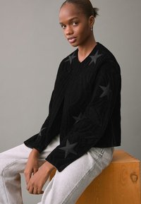 Chaqueta negra texturizada con un diseño acolchado y acentos de estrellas, combinada con jeans de color gris claro. El modelo está sentado sobre una superficie de madera.