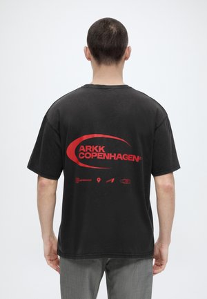 Άνδρας με κοντά μαλλιά που φοράει μαύρο μπλουζάκι ARKK Copenhagen με μεγάλο κόκκινο λογότυπο στην πλάτη, στέκεται μπροστά σε απλό φόντο.