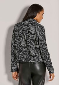 Frau mit langen dunklen Haaren, die eine Jacke mit schwarzem und weißem Paisleymuster sowie schwarze Lederen besitzt, von hinten gegen einen einfarbigen Hintergrund gerichtet.