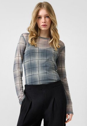 Junge Frau mit langen blonden Haaren, die ein graues kariertes Langarmshirt und schwarze hoch taillierte Hosen trägt, steht vor einem schlichten Hintergrund.