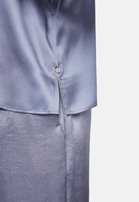 Zwei Kleidungsstücke: eine seidige, hellviolette Bluse mit einem Knopf und eine Paar strukturierte, glänzende hellviolette Hosen.