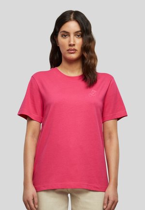 Femme aux longs cheveux ondulés foncés portant un t-shirt rose vif à manches courtes avec un petit logo cerise sur la poitrine gauche, sur un fond gris.