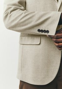 Blazer texturizado en color beige con un corte ajustado, cuatro botones negros en el puño y un bolsillo en el pecho.
