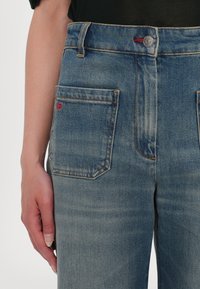 Jeans in denim blu chiaro con vita alta, due tasche posteriori e dettagli di cucitura rossa sulla tasca sinistra. Tessuto morbido e taglio classico.